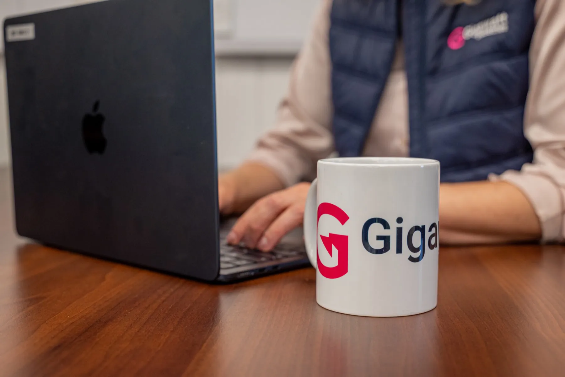 A branded Gigawatt mug on a desk beside a laptop, with a colleague working behind it